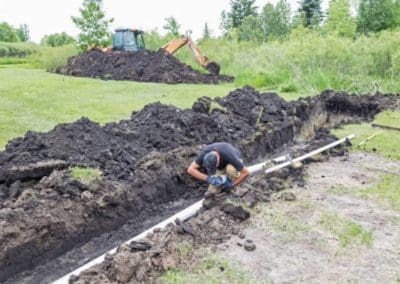 tech installing septic system plumbing with an excavator in the background